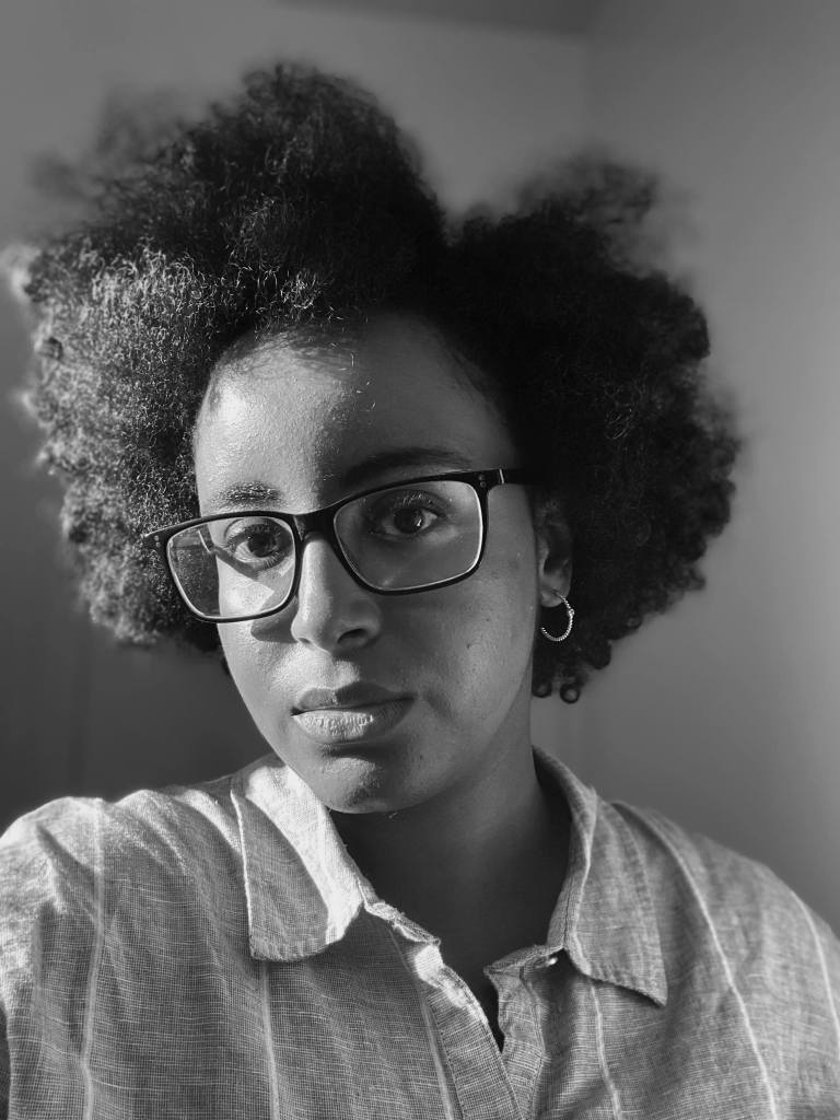 Black and white photo of a young black woman with black curly hair, collared shirt and black square framed glasses.